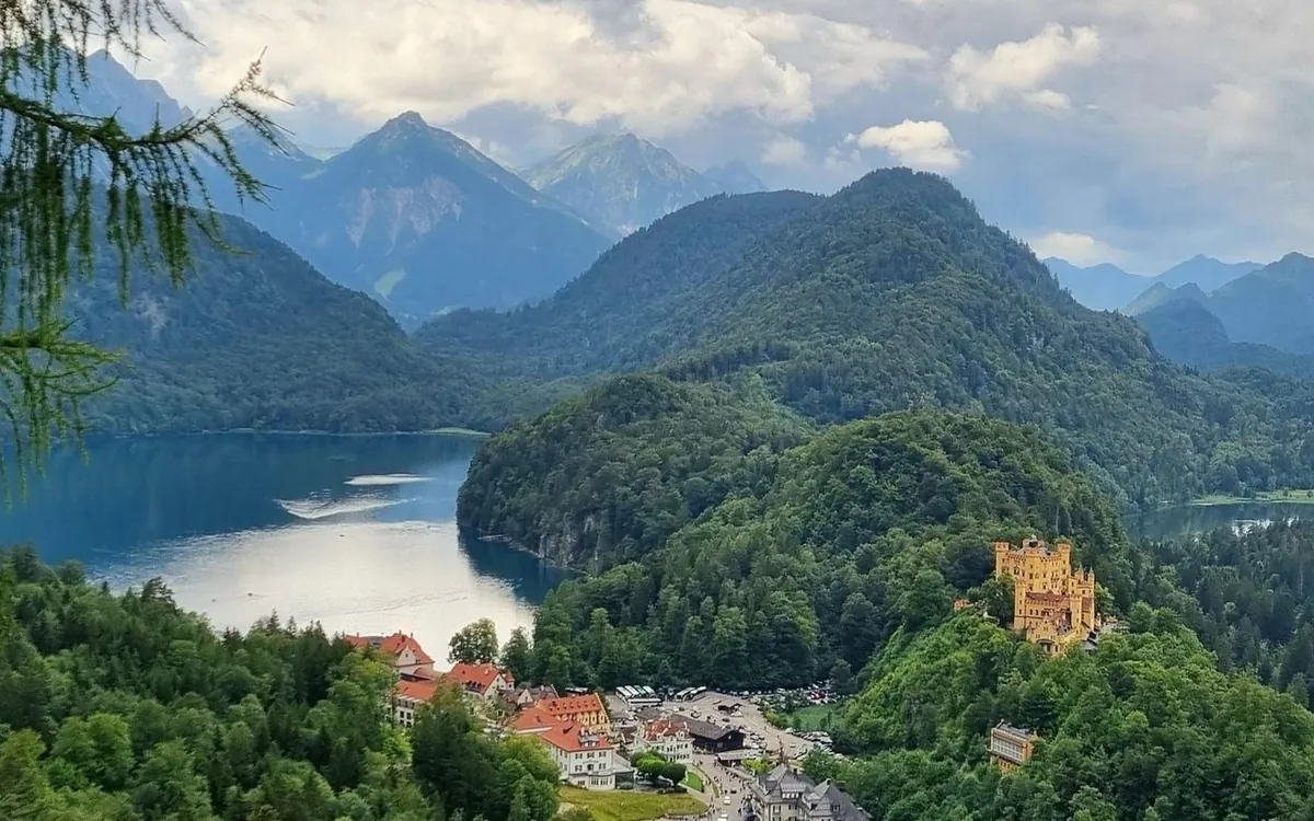 Der Alpsee und das gelbe Schloss Hohenschwangau aus der Höhe gesehen, dahinter Berge