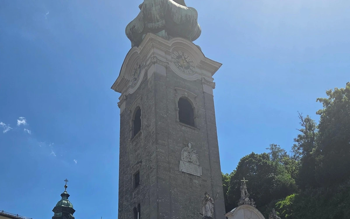 Una alta torre de iglesia de piedra con esfera de reloj y cúpula bulbosa verde contra un cielo azul, con estatuas a sus pies