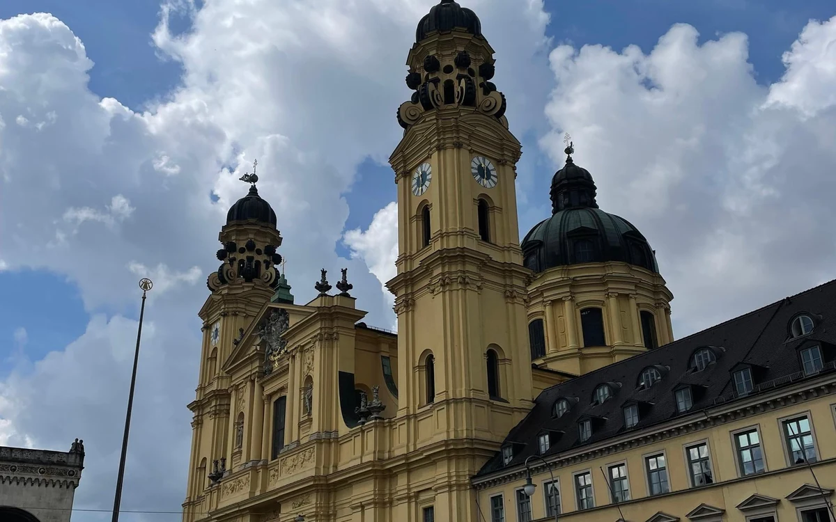 Una chiesa barocca gialla con due torri dell'orologio e una cupola verde, accanto a un lungo edificio dal tetto grigio