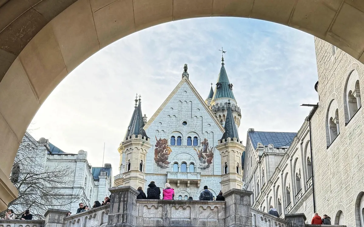 The pale gabled front of Neuschwanstein seen through a stone arch, with visitors at the balustrade