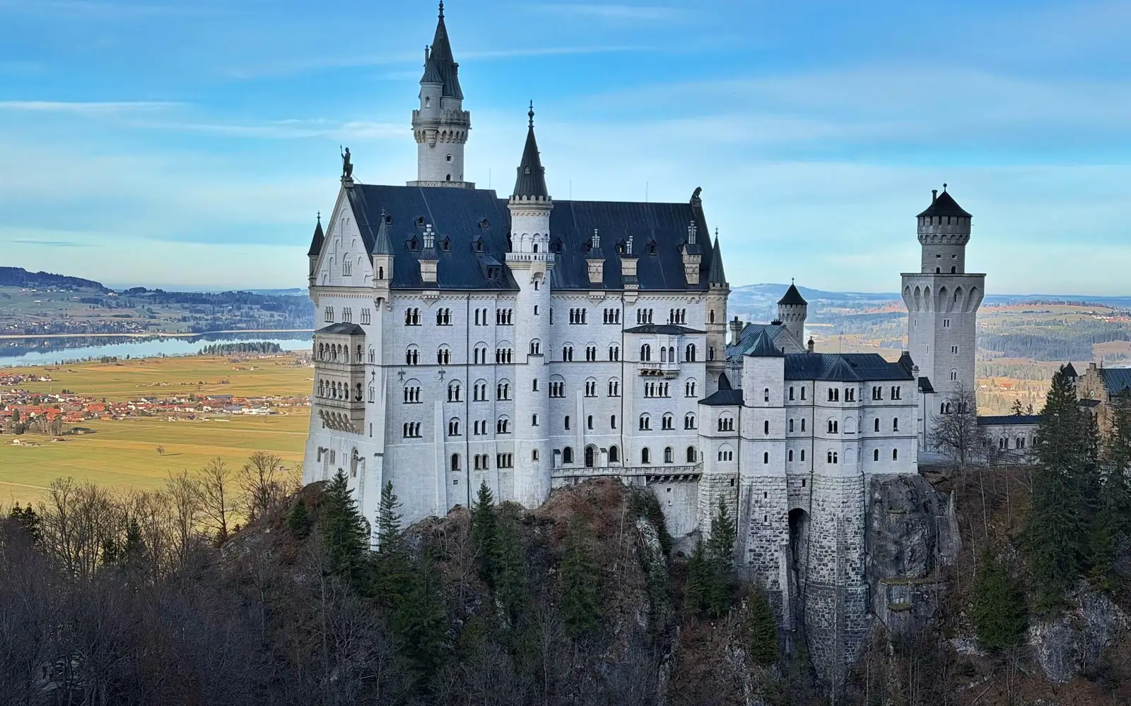 Neuschwanstein Castle auf seinem Felsgrat über dem Wald, dahinter ein See und Felder