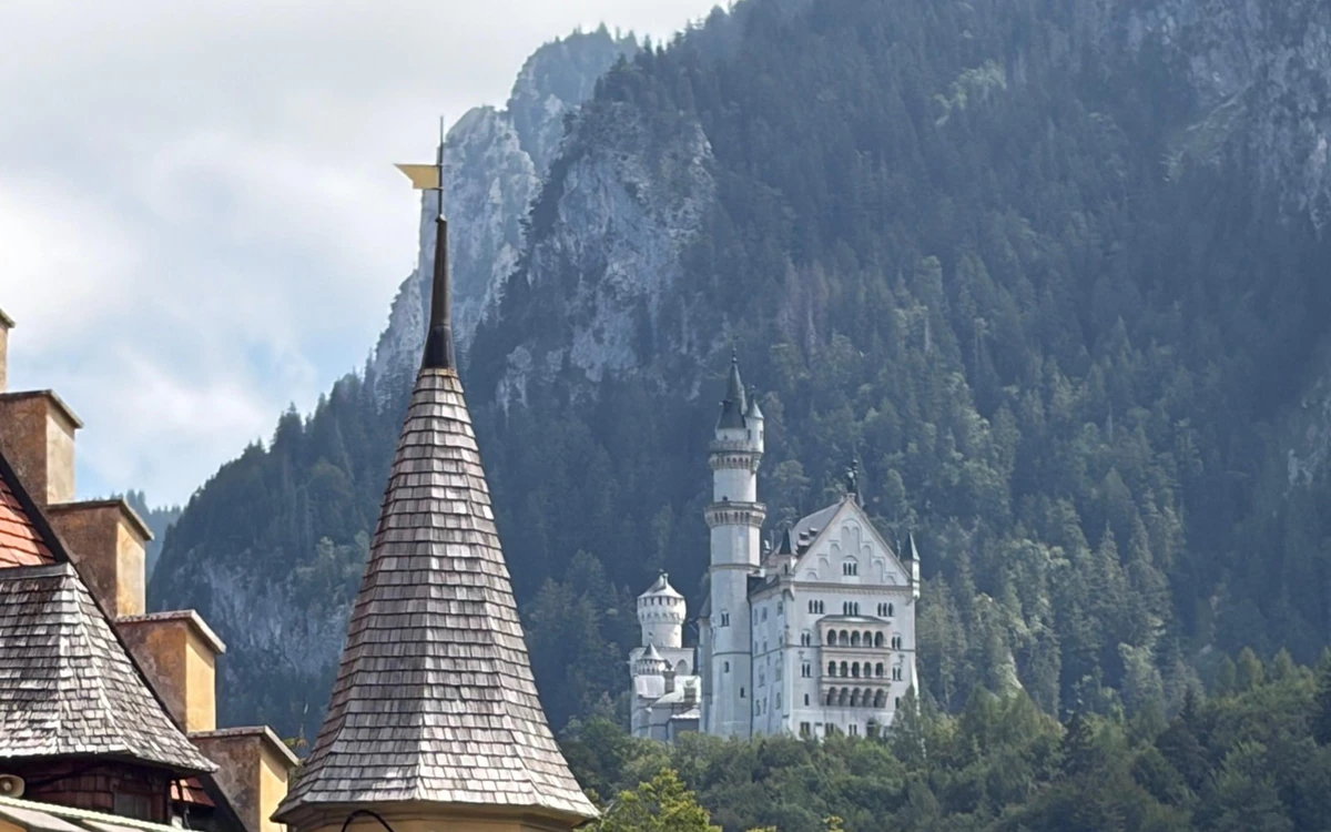 A shingled tower roof in the foreground and Neuschwanstein below a steep forested mountainside