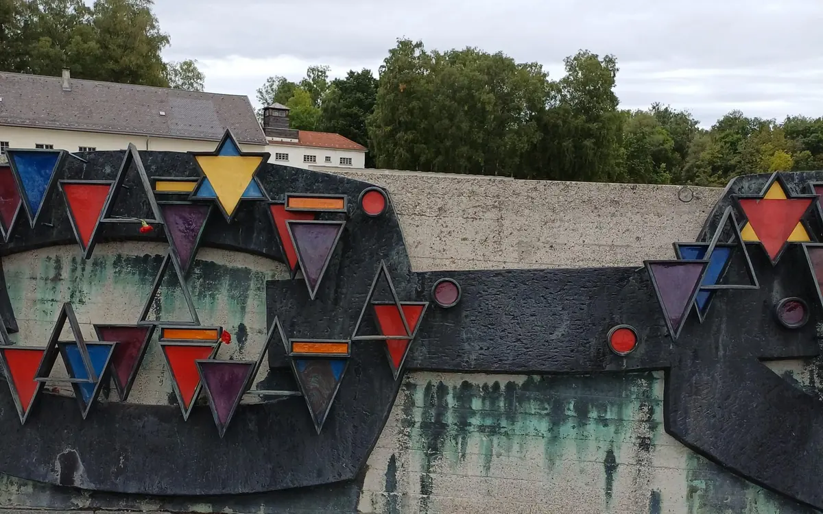 Un bassorilievo commemorativo in metallo scuro con triangoli colorati davanti a un lungo edificio bianco