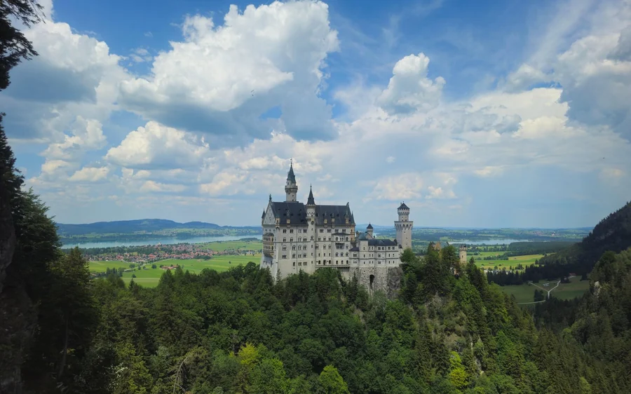 Neuschwanstein Castle über einem bewaldeten Tal, in der Ferne Seen und Felder