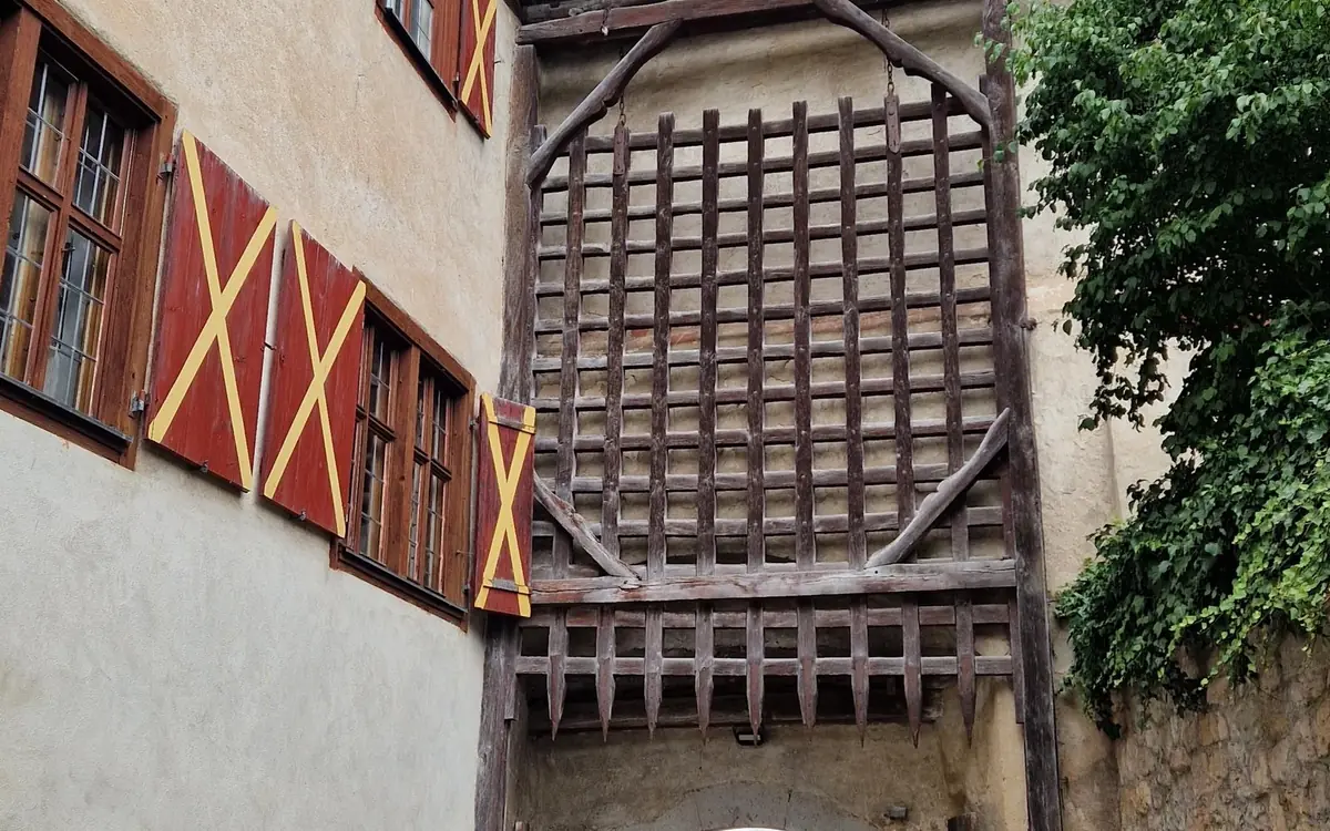 A wooden portcullis raised above a gateway, beside red shutters with yellow crosses